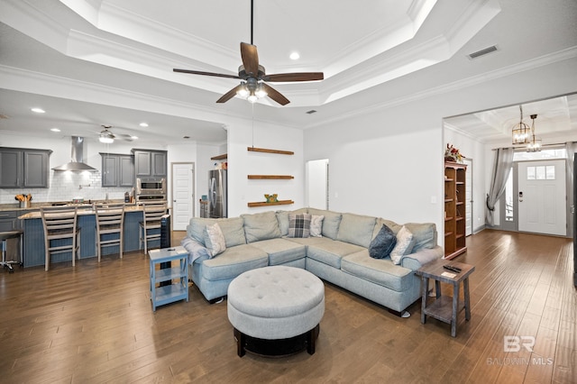 living room featuring dark wood-type flooring, ornamental molding, a raised ceiling, and ceiling fan with notable chandelier