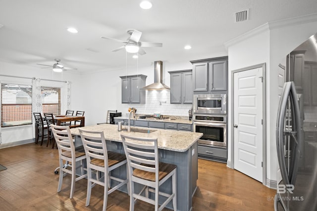 kitchen featuring a kitchen breakfast bar, stainless steel appliances, light stone counters, an island with sink, and wall chimney exhaust hood