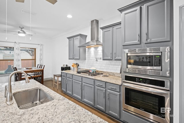 kitchen with wall chimney range hood, sink, gray cabinets, appliances with stainless steel finishes, and light stone countertops