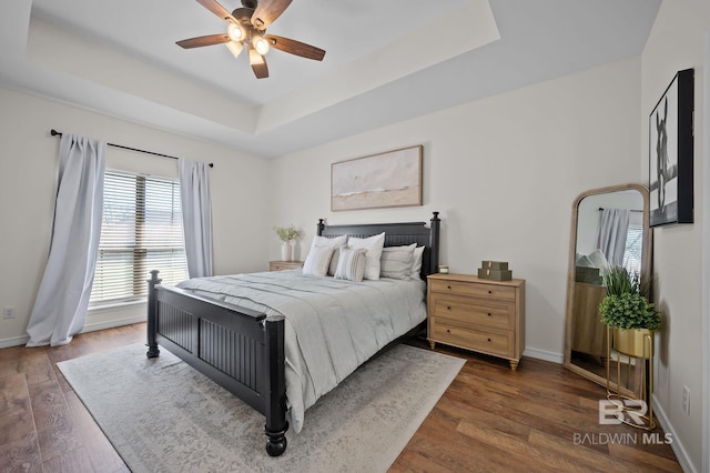 bedroom with baseboards, dark wood-type flooring, a tray ceiling, and a ceiling fan