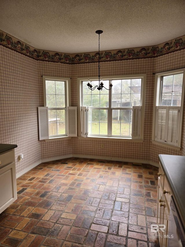 unfurnished dining area with brick floor, a notable chandelier, a textured ceiling, baseboards, and wallpapered walls