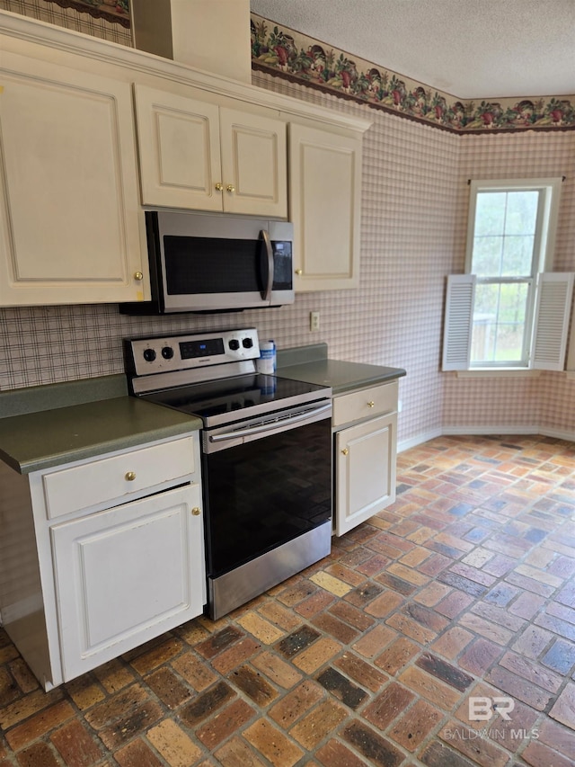 kitchen featuring brick floor, cream cabinetry, dark countertops, appliances with stainless steel finishes, and wallpapered walls