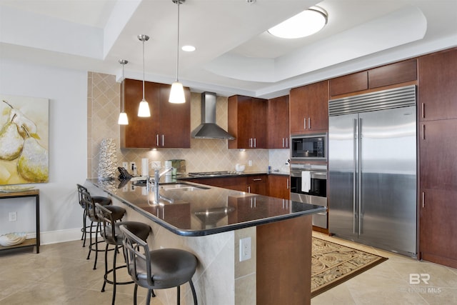 kitchen with built in appliances, a peninsula, a sink, wall chimney range hood, and a tray ceiling