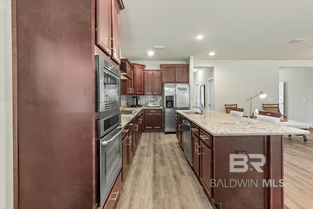 kitchen with appliances with stainless steel finishes, a sink, light wood finished floors, light stone counters, and an island with sink