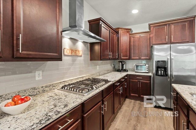kitchen featuring stainless steel appliances, wall chimney exhaust hood, light wood-style flooring, decorative backsplash, and light stone counters