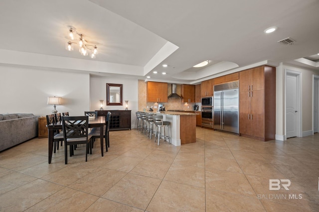 kitchen with brown cabinetry, wall chimney exhaust hood, a raised ceiling, and built in appliances