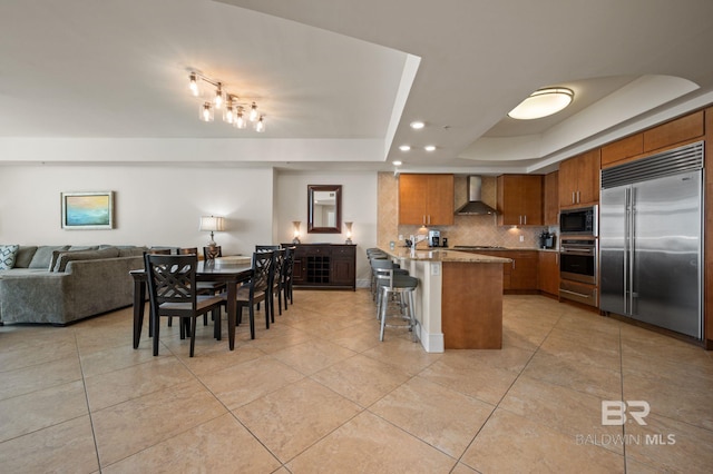 kitchen featuring brown cabinets, a raised ceiling, open floor plan, built in appliances, and wall chimney exhaust hood