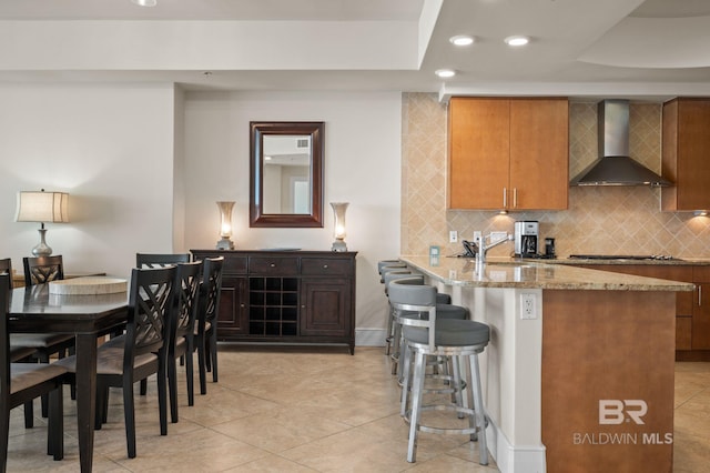 kitchen featuring recessed lighting, a breakfast bar, light stone countertops, wall chimney exhaust hood, and tasteful backsplash