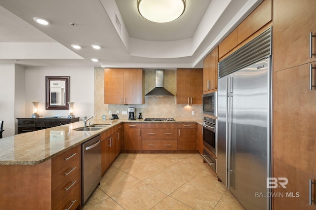 kitchen with wall chimney exhaust hood, built in appliances, a peninsula, light stone countertops, and a sink