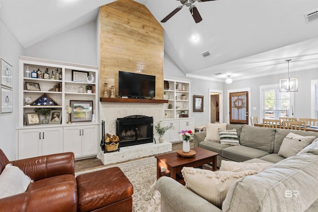 living room featuring lofted ceiling, a fireplace, built in features, ornamental molding, and ceiling fan with notable chandelier