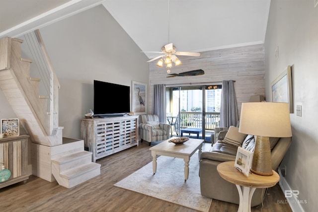 living area featuring wooden walls, ceiling fan, stairway, wood finished floors, and high vaulted ceiling