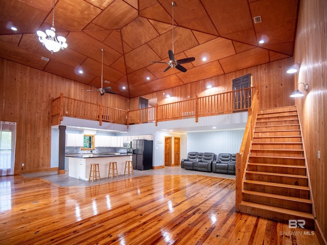unfurnished living room featuring wood walls, stairs, a towering ceiling, a ceiling fan, and light wood-type flooring
