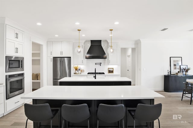 kitchen with premium range hood, hanging light fixtures, stainless steel appliances, an island with sink, and white cabinets