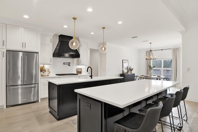 kitchen featuring pendant lighting, high end fridge, an island with sink, white cabinets, and custom exhaust hood