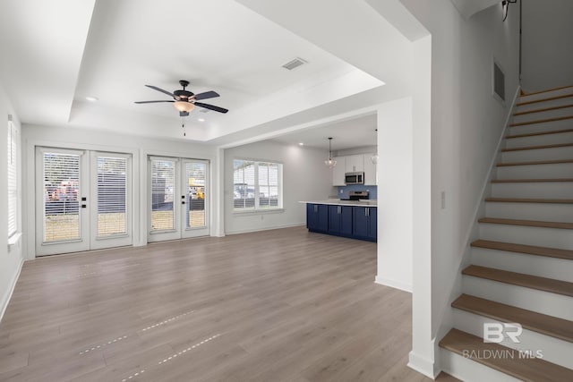 unfurnished living room featuring visible vents, ceiling fan, stairway, a tray ceiling, and light wood-type flooring