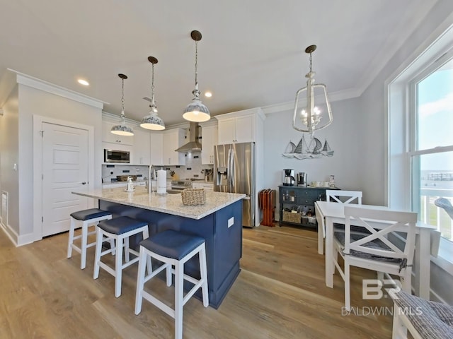 kitchen featuring white cabinetry, hanging light fixtures, stainless steel appliances, a center island with sink, and wall chimney range hood