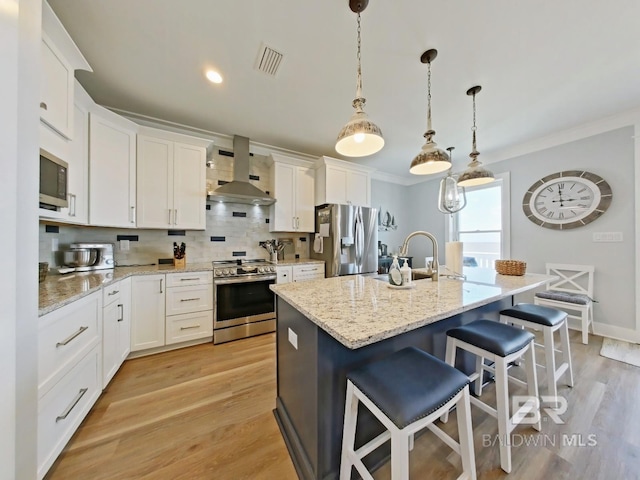 kitchen featuring wall chimney range hood, a kitchen island with sink, white cabinetry, hanging light fixtures, and stainless steel appliances