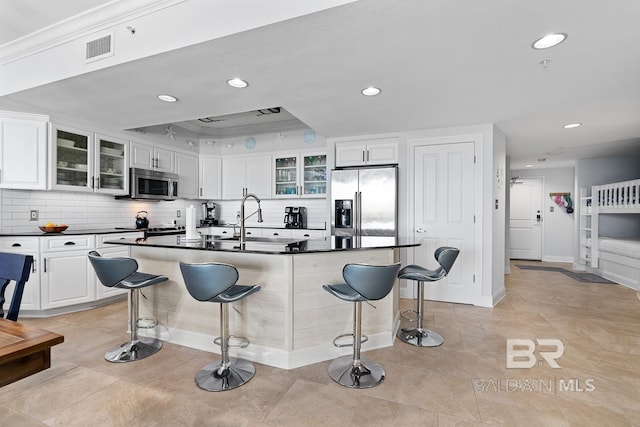 kitchen with a kitchen bar, a center island with sink, stainless steel appliances, white cabinetry, and light tile floors