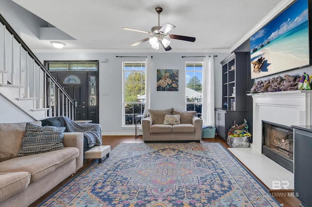 living area with a ceiling fan, a textured ceiling, a fireplace, crown molding, and stairs