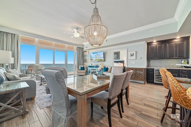 dining area with wine cooler, light wood-style flooring, ornamental molding, wet bar, and ceiling fan with notable chandelier