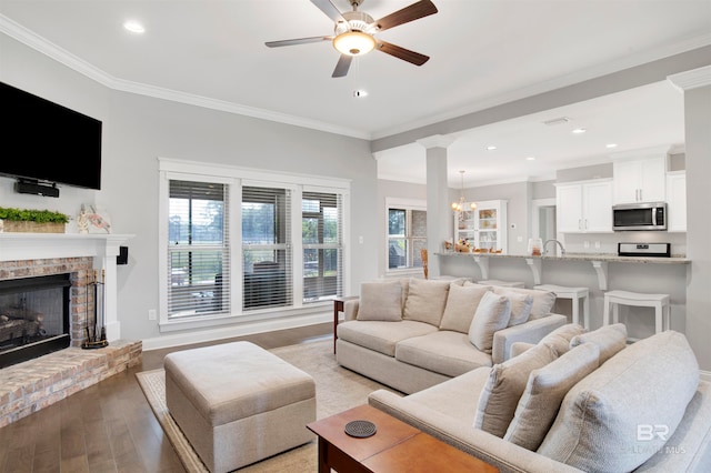 living room with a brick fireplace, ceiling fan with notable chandelier, light hardwood / wood-style flooring, ornamental molding, and decorative columns