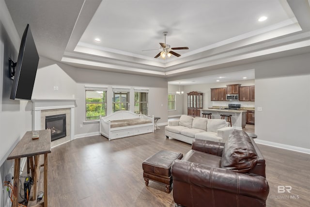 living room featuring a tile fireplace, ceiling fan with notable chandelier, a raised ceiling, dark hardwood / wood-style floors, and ornamental molding