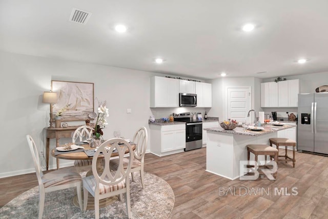 kitchen with white cabinetry, appliances with stainless steel finishes, light wood-type flooring, light stone countertops, and an island with sink