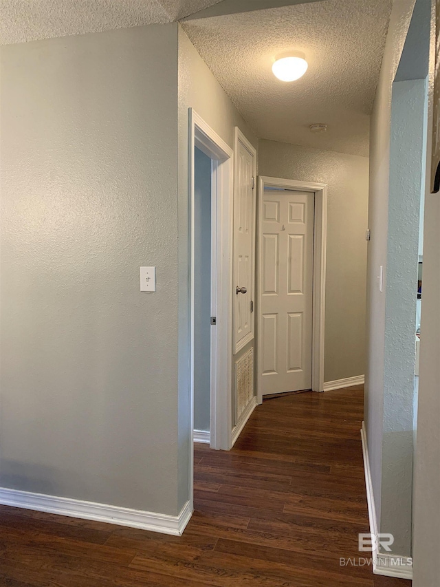 hall with dark wood-type flooring, a textured ceiling, a textured wall, and baseboards