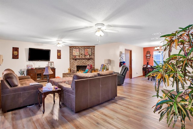 living room featuring a textured ceiling, light hardwood / wood-style flooring, a brick fireplace, and ceiling fan with notable chandelier