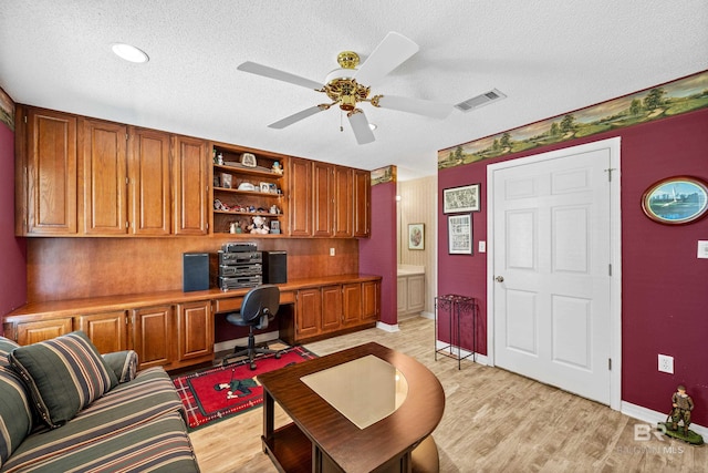 office area with built in desk, visible vents, light wood-style flooring, ceiling fan, and a textured ceiling