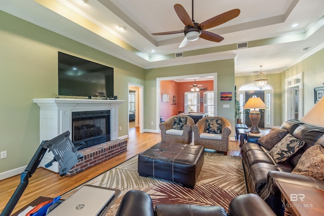 living area featuring visible vents, a brick fireplace, crown molding, a tray ceiling, and wood finished floors