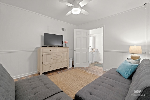 living area with visible vents, a ceiling fan, ornamental molding, wainscoting, and light wood-type flooring