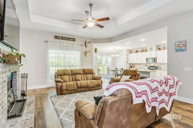 living room with ceiling fan with notable chandelier, hardwood / wood-style flooring, a tray ceiling, and a brick fireplace