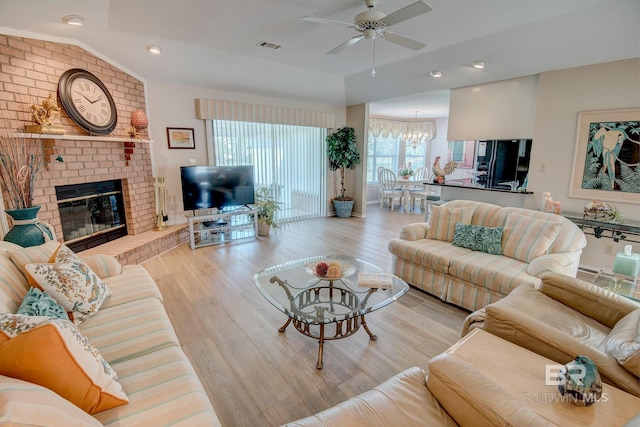 living room featuring lofted ceiling, light hardwood / wood-style flooring, ceiling fan with notable chandelier, a fireplace, and brick wall