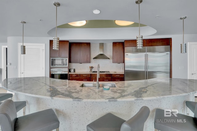 kitchen with wall chimney range hood, decorative backsplash, sink, built in appliances, and hanging light fixtures