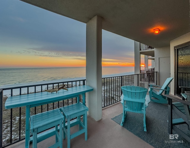 balcony at dusk featuring a water view and a beach view