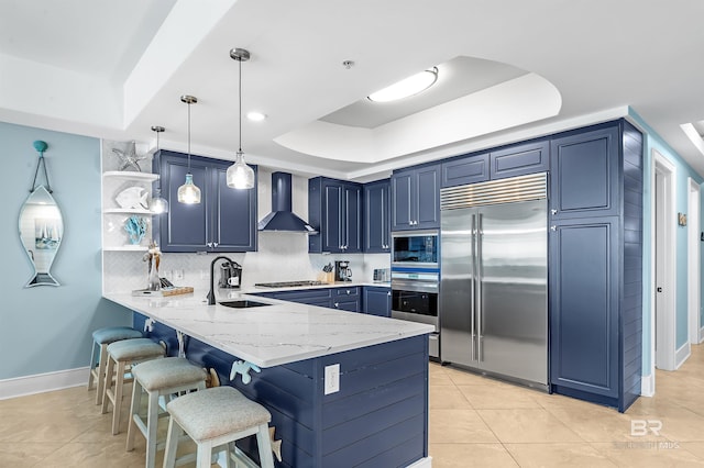 kitchen with a tray ceiling, a sink, wall chimney range hood, built in appliances, and a peninsula