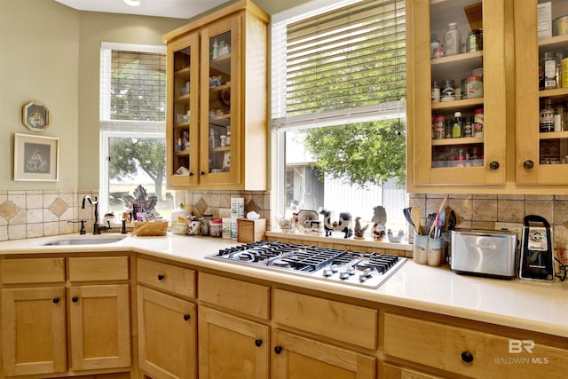kitchen with stainless steel gas cooktop, a sink, light countertops, backsplash, and glass insert cabinets