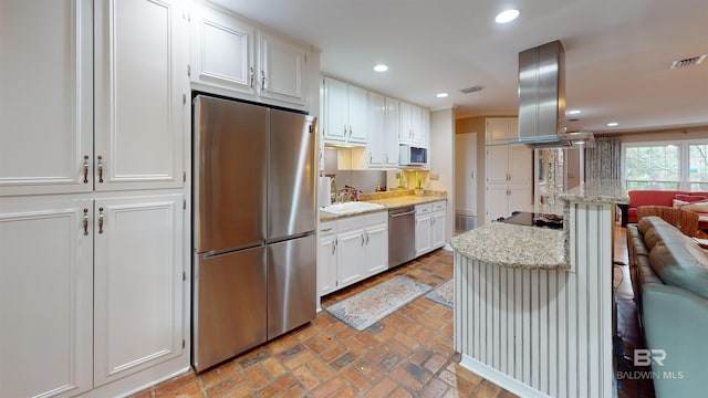 kitchen featuring light stone countertops, appliances with stainless steel finishes, island range hood, sink, and white cabinets