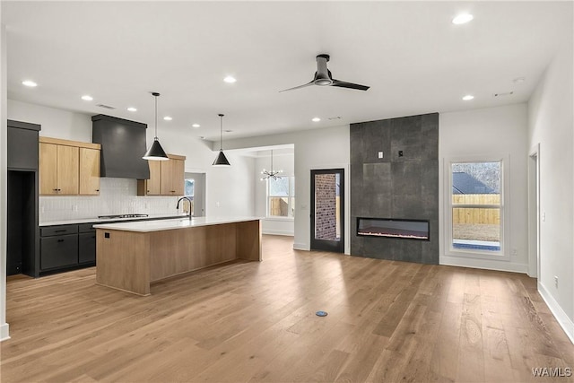 kitchen with decorative light fixtures, a large fireplace, custom range hood, a center island with sink, and light hardwood / wood-style flooring