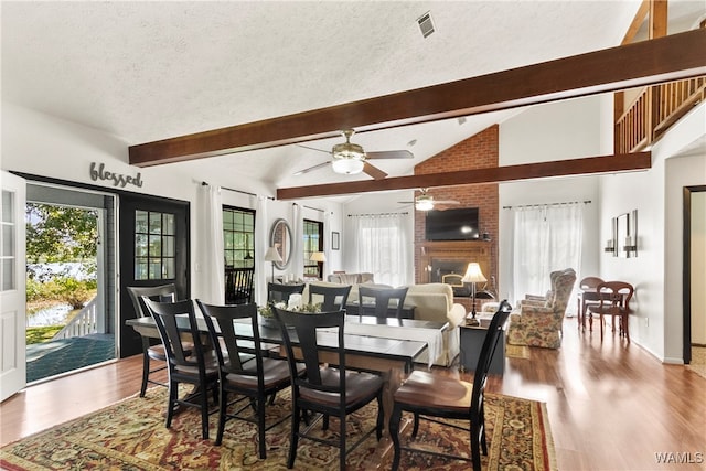 dining room with ceiling fan, a brick fireplace, lofted ceiling with beams, wood-type flooring, and a textured ceiling