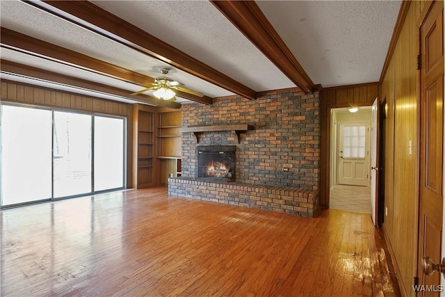 unfurnished living room featuring wood walls, beam ceiling, a brick fireplace, a textured ceiling, and light wood-type flooring