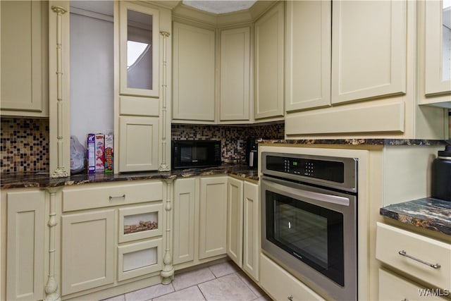 kitchen featuring backsplash, stainless steel oven, cream cabinets, light tile patterned floors, and dark stone countertops