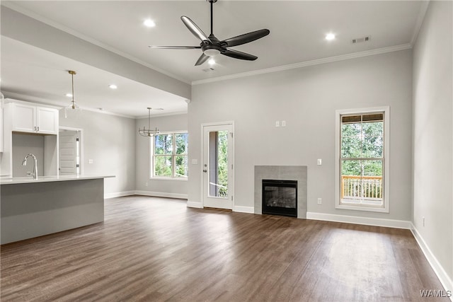 unfurnished living room with a tile fireplace, ceiling fan with notable chandelier, sink, crown molding, and dark hardwood / wood-style floors