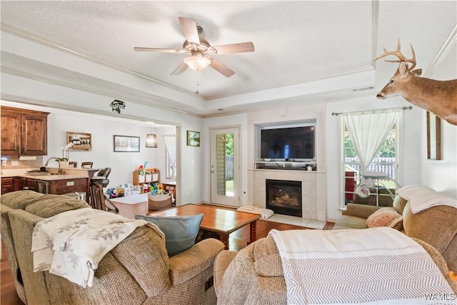 living area featuring crown molding, a fireplace, ceiling fan, a raised ceiling, and light wood-style flooring