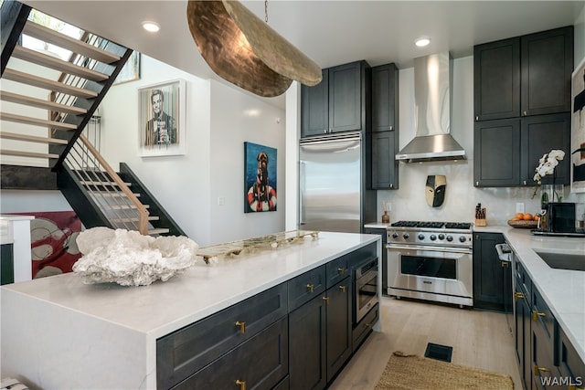 kitchen featuring wall chimney exhaust hood, hanging light fixtures, built in appliances, light hardwood / wood-style floors, and a kitchen island