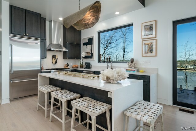 kitchen with a breakfast bar, wall chimney exhaust hood, hanging light fixtures, and stainless steel built in refrigerator