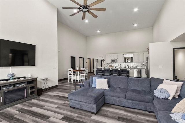 living area with recessed lighting, a towering ceiling, a ceiling fan, wood finished floors, and baseboards