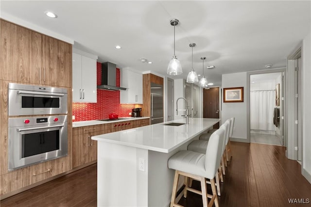 kitchen with appliances with stainless steel finishes, an island with sink, sink, white cabinets, and wall chimney range hood