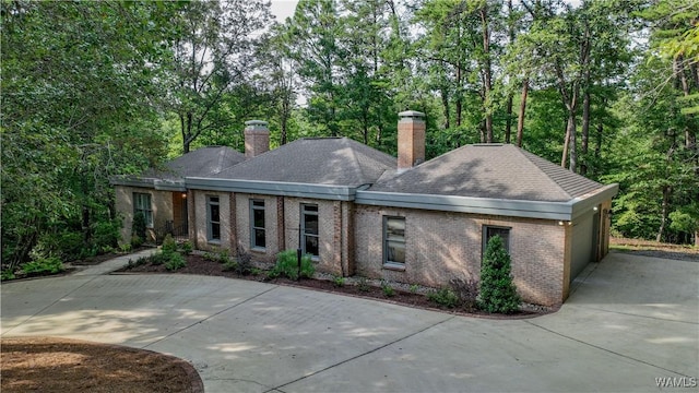 rear view of property featuring brick siding, an attached garage, a shingled roof, a chimney, and driveway